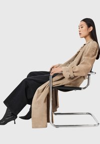 Μπεζ σουέντ καμπαρντίνα με φαρδιά μανίκια, σε συνδυασμό με μαύρο τζιν παντελόνι φαρδιάς γραμμής και μυτερές μπότες αστραγάλου, καθισμένη σε λεπτή καρέκλα από χρώμιο.