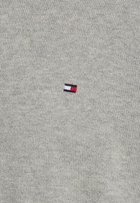 Pulover gri din tricot, cu o textură moale, având un mic logo brodat în roșu, alb și albastru pe partea stângă a pieptului.