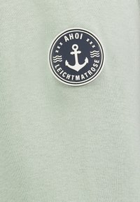Lichtgroene stof met een gestructureerde afwerking, met een ronde marineblauwe patch met een anker en de tekst "AHOI LEICHTMATROSE."