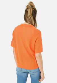 Pull à col rabattu à manches courtes en tricot orange vif. Présente une texture lisse et un petit détail accentué près du col. Associé à un jean bleu.