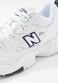 Bílá kožená teniska s námořnicky modrými doplňky, výrazným logem „N“, texturovanými panely a plochými tkaničkami na polstrované podrážce.