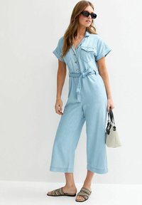 Une combinaison en denim bleu clair avec des manches courtes, un devant bouton, une taille à nouer et des jambes larges. Portée avec des sandales rayées et un petit sac à main.