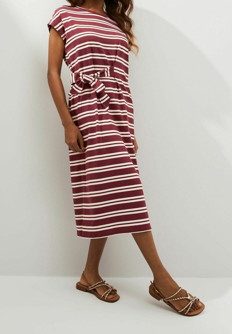 Femme portant une robe midi rayée à manches courtes avec une ceinture nouée à la taille, associée à des sandales plates à brides tressées.