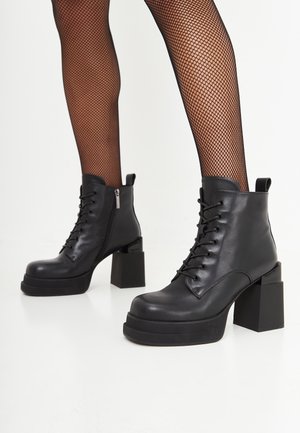 Jambes portant des bas résille noirs et des bottines noires épaisses à plateforme lacées à la cheville avec talons blocs sur fond blanc.