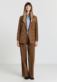 Blazer doppiopetto in velluto a coste marrone e pantaloni a gamba larga abbinati, abbinati a una camicia azzurra con bottoni. Texture liscia, vestibilità su misura.