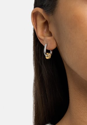 Srebrna kolczyk w kształcie koła z małym złotym pierścieniem ozdobionym kryształkami, noszony na lewym uchu osoby na białym tle.