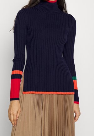 Femme portant un pull à col roulé côtelé bleu marine avec des bandes rouges, orange et vertes sur les manches, associé à une jupe plissée beige.
