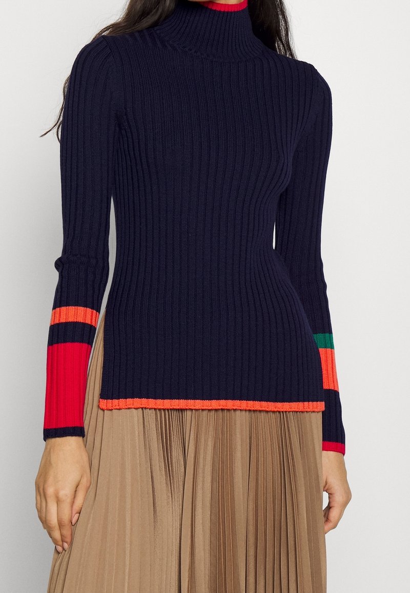 Femme portant un pull à col roulé côtelé bleu marine avec des bandes rouges, orange et vertes sur les manches, associé à une jupe plissée beige.