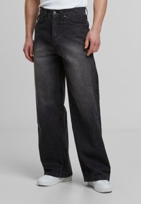 Jeans de mezclilla anchos en negro con un acabado desgastado, cinco bolsillos, cierre de botón metálico y costuras en contraste, combinados con zapatillas blancas.