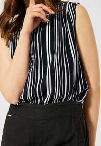 Kvinna som bär en ärmlös svart och vit vertikalt randig topp instoppad i höga svarta byxor, med handen nära ansiktet.