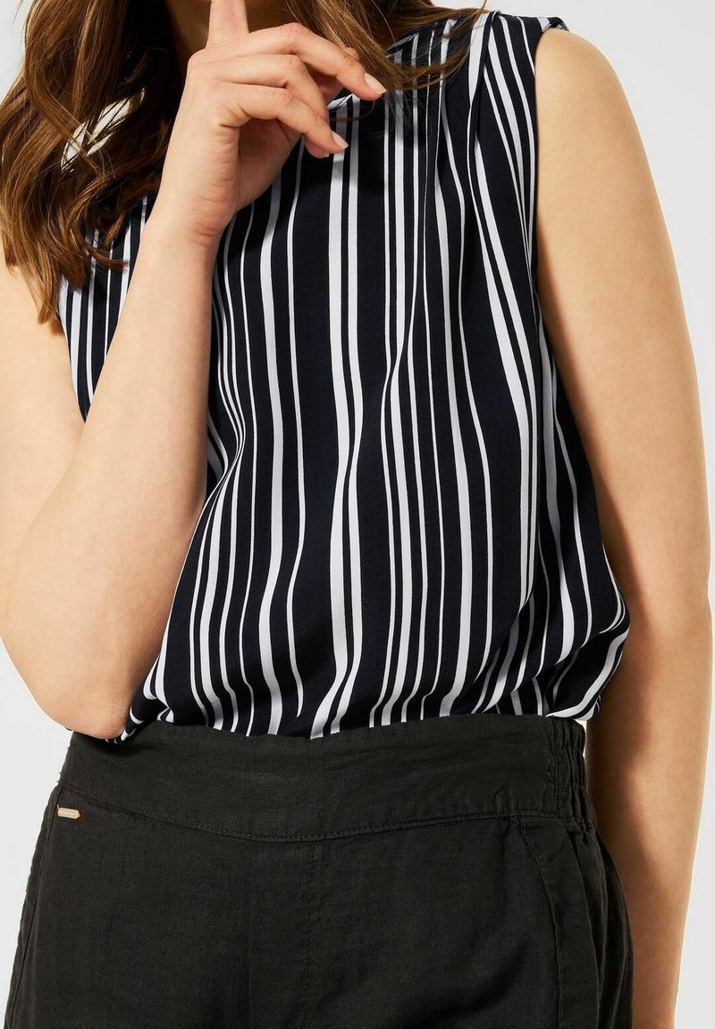 Kvinna som bär en ärmlös svart och vit vertikalt randig topp instoppad i höga svarta byxor, med handen nära ansiktet.