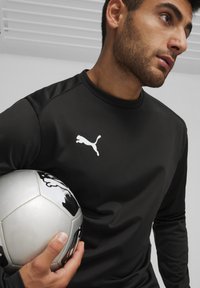 Zwart sporthemd met lange mouwen en een wit Puma-logo op de borst, vastgehouden met een hand, naast een zilveren en zwart voetbalt. 