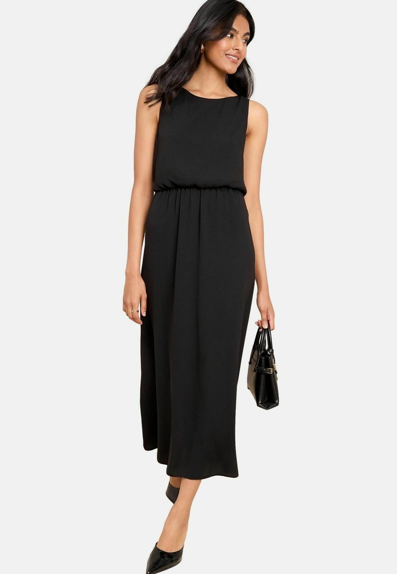 Schwarzes ärmelloses Midikleid mit rundem Ausschnitt, geraffter Taille und fließender Silhouette. Kombiniert mit schwarzen spitzen Absatzschuhen und einer Handtasche.
