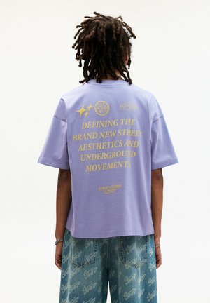 Lilla oversized t-shirt med guldtekst på ryggen, prydet med grafiske logoer. Fremstillet af bomuld, med korte ærmer og afslappet pasform.
