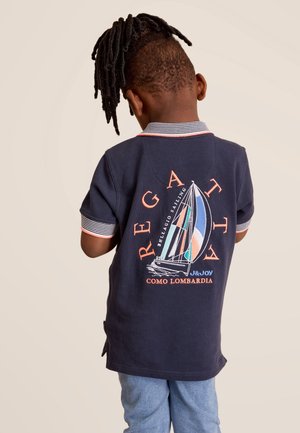 Kind mit Dreadlocks trägt ein dunkelblaues Polohemd mit buntem Segelboot-Motiv und Text auf dem Rücken, kombiniert mit hellblauen Hosen.