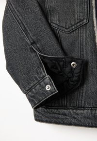 Manga de chaqueta de mezclilla gris oscura con forro interior negro, botón a presión plateado y detalles de costura en contraste.