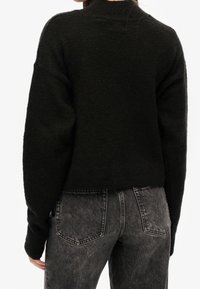 Czarny sweter z dzianiny o wysokim kołnierzu, w oversize'owym fasonie i teksturowanej tkaninie, zestawiony z wyblakłymi szarymi dżinsami z tylnymi kieszeniami.