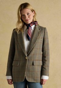 Blazer s vzorom houndstooth v zeleno-hnedo-červenej farbe. Obsahuje jeden zlatý gombík, strih na mieru a dve predné vrecká. Nosené na bielu košeľu.