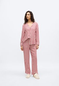 Γυναίκα στέκεται φορώντας κόκκινη και λευκή πιτζάμα με καρδιές και μπεζ παντόφλες σε απλό φόντο.