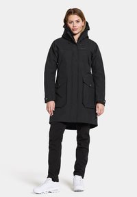 Schwarzer Parka mit Kapuze, Reißverschluss vorne und zwei großen Taschen. Verfügt über einen geraden Schnitt und strapazierfähigen Stoff. Wird über schwarze Hosen und weißen Turnschuhen getragen.