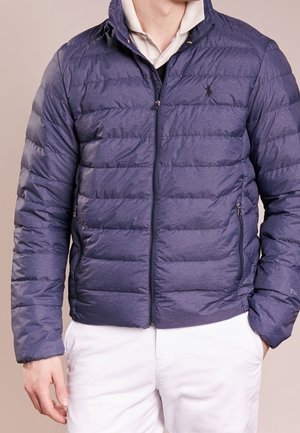 Homme portant une doudoune matelassée bleu foncé sur un polo beige clair avec un pantalon blanc, les mains reposant calmement sur les côtés.
