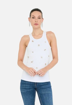 La modella si erge con sicurezza, indossando un top bianco senza maniche con decorazioni e abbinato a jeans in denim blu.