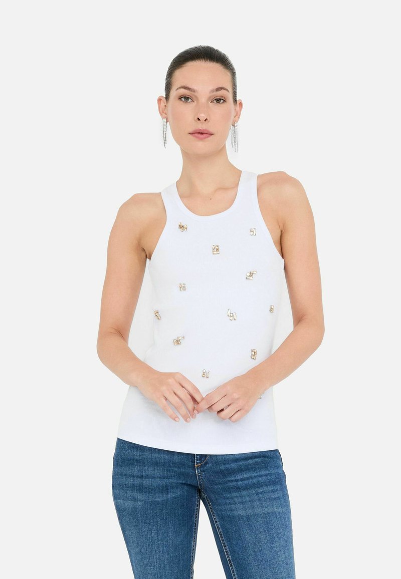 Model staat zelfverzekerd, gekleed in een mouwloos wit topje met decoratieve versieringen, gecombineerd met blauwe denim jeans.