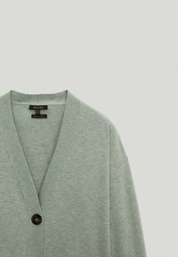 Jasnozielony kardigan z dekoltem w serek, długimi rękawami i jednym czarnym guzikiem. Miękka tekstura, minimalistyczny design i brak widocznych wzorów.