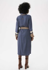 Personne aux cheveux bouclés portant une robe à manches longues bleu et beige à motifs et des bottines à talons marron, se tenant devant un fond blanc.