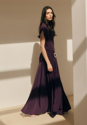 Mujer con un vestido fluido de color púrpura oscuro con un cinturón adornado con joyas, de pie bajo una suave luz solar que proyecta sombras sobre una pared beige.