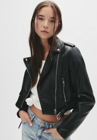 Zwarte cropped leren motorjack met ritsen, schouderepauletten en een gespdetail. Draagt over een witte top en lichtblauwe jeans.