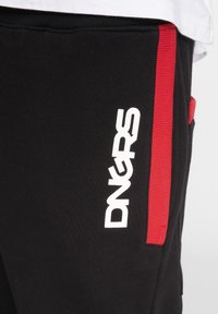 Schwarze Jogginghose mit einem weißen "DNGRS"-Logo, ausgestattet mit einem roten Seitenstreifen und einem strukturierten Bund, der durch eine Taschenapplikation hervorgehoben wird.