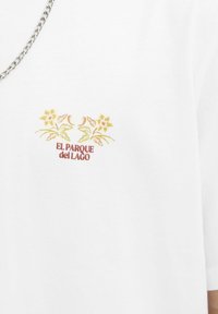 Hvid T-shirt med korte ærmer, prydet med floral broderi i gult og brunt over teksten "EL PARQUE del LAGO."