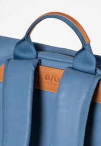 Zaino blu con materiale testurizzato, dettagli in pelle marrone e manico imbottito. L'etichetta del marchio è impressa sulla toppa in pelle.