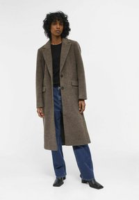 Lange, bruine wollen jas met een ingesneden kraag en twee voorzakken, gecombineerd met donkere jeans en zwarte laarzen met vierkante neuzen.