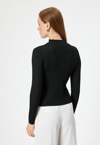 Schwarzes, geripptes Langarmoberteil mit hohem Kragen und taillierter Silhouette. Sichtbares Texturmuster, kombiniert mit weißen Hosen und schlichten Ohrringen.