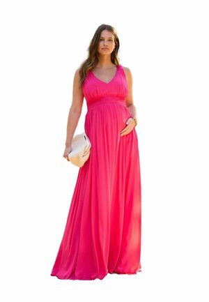 Femme portant une longue robe maxi rose sans manches, tenant une pochette beige en tissu tressé, avec des cheveux ondulés lâches et des bijoux minimalistes.