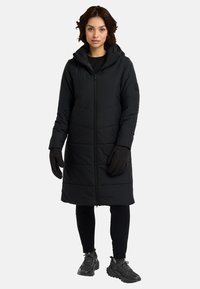 Lungo cappotto imbottito nero con cappuccio, chiusura frontale con zip e dettagli a coste. Indossato sopra leggings neri e guanti, abbinato a calzature scure.