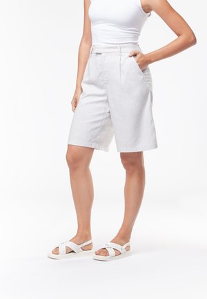 Donna che indossa un top bianco senza maniche, pantaloncini bianchi a vita alta fino al ginocchio e sandali bianchi con suola piatta, in piedi con una mano in tasca.