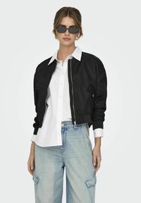 Veste bomber noire avec fermeture éclair et poches latérales, portée sur une chemise blanche à boutons et un jean baggy bleu clair. Lunettes de soleil noires portées.