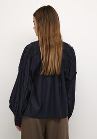 Marineblaue Bluse mit langen, luftigen Ärmeln und strukturiertem Besatz entlang der Schultern. Weicher Stoff und lockere Passform, kombiniert mit braunen Hosen.
