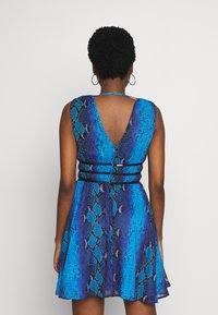 Femme portant une robe sans manches à imprimé serpent bleu et violet avec un dos en V profond et des détails de ceinture noire, se tenant devant un fond uni.