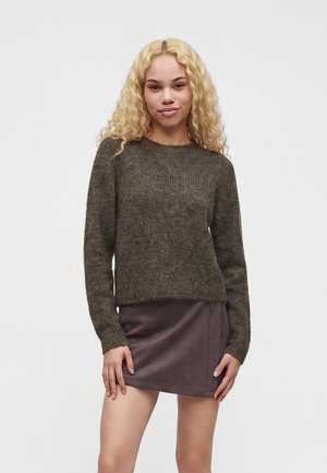 Jeune femme aux cheveux blonds bouclés portant un pull en laine marron foncé et une mini-jupe marron, debout devant un fond clair uni.