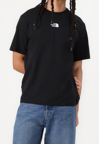 Black cotton t-shirt featuring a small white logo on the chest, with short sleeves and a relaxed fit, paired with blue denim jeans.