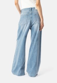 Osoba oblečená v svetlomodrých širokých džínsach a bielej blúzke, zobrazená zozadu na čistom bielom pozadí.