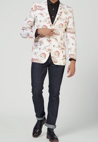 Witte blazer met kerstthema-illustraties van de kerstman, hulst en linten. Gecombineerd met donkere jeans en zwarte schoenen met blauwe accenten.
