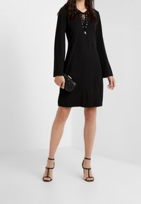 Czarna długa sukienka z długim rękawem i dekoltem wiązanym w koronkę, gładka tekstura i luźny krój; zestawiona z czarnymi sandałami na obcasie i małą czarną torebką.