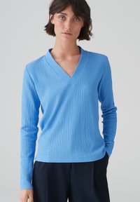 Helles blaues LangARM-Shirt mit V-Ausschnitt, strukturiertem Stoff, figurbetontem Schnitt und dezenten Rippdetails, kombiniert mit dunkelblauen Hosen.
