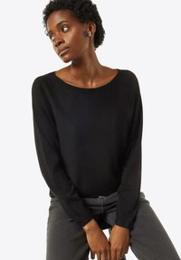 Schwarzer Strickpullover mit weitem Ausschnitt und langen Ärmeln, getragen mit grauen Jeans. Der Hintergrund ist neutral, um das Kleidungsstück hervorzuheben.