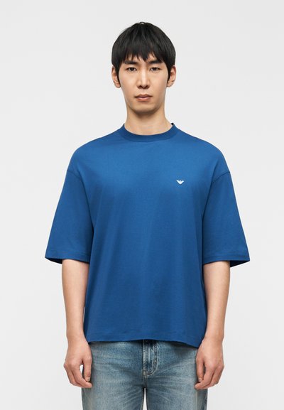 Μπλε βαμβακερό t-shirt με στρογγυλή λαιμόκοψη και κοντά μανίκια, με ένα μικρό λευκό λογότυπο στο στήθος. Συνδυάζεται με μπλε τζιν παντελόνι.